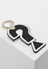 Zwart-witte sleutelhanger in de vorm van een vraagteken, gemaakt van leer, met een zilveren ring en een klein labeltje met "GUESS" eraan bevestigd.