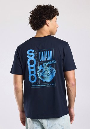 Jonge man met krullend haar draagt een zwart T-shirt met blauwe grafische tekst "SOHO UMAMI" en een kom ramen met eetstokjes op de achterkant.