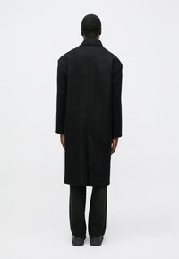 Lungo cappotto nero in lana con taglio dritto, spalle larghe e cucitura centrale sulla schiena, abbinato a pantaloni scuri ampi e scarpe nere.