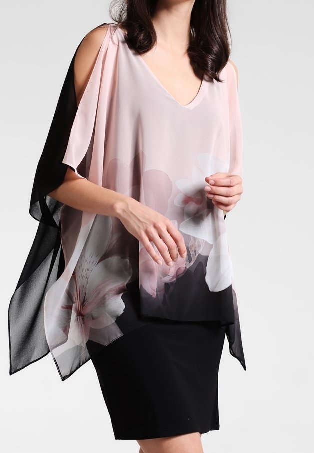 Donna che indossa un top floreale trasparente rosa e nero fluente su una gonna nera aderente, davanti a uno sfondo chiaro e semplice.