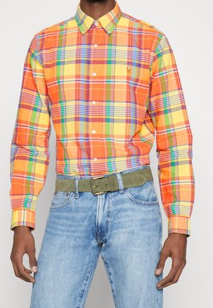 Homme portant une chemise à carreaux multicolores orange, jaune et verte, avec un jean bleu et une ceinture vert olive.