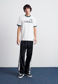 Fiatal férfi áll fehér Adidas pólóban zöld díszítéssel, fekete Adidas melegítőnadrágban fehér csíkokkal, és fekete-fehér sportcipőben.