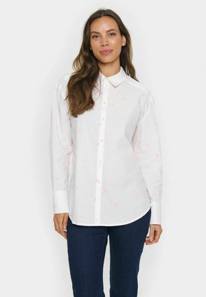 Femme aux longs cheveux bruns portant une chemise blanche à boutons ornée de petits motifs en forme de cœur roses et un jean bleu foncé, se tenant devant un fond uni.