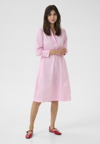 Šviesiai rožinė suknelė su apykakle, trijų ketvirčių rankovėmis ir laisvu siluetu. Derinama su raudonomis butais, turinčiais dekoratyvius akcentus.