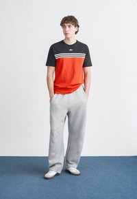 T-shirt vermelha e preta com riscas brancas, combinada com calças de treino cinzentas. Material de algodão, corte descontraído, modelo em pé contra um fundo simples.