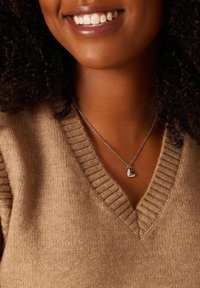 Zilveren hart hanger ketting op een fijne ketting, gedragen met een beige gebreide trui met een V-hals en geribbelde textuur.