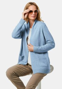 Hellblauer Cardigan aus weichem Strickstoff, offenes Design mit gerippten Kanten, ausgestattet mit zwei vorderen Taschen. Kombiniert mit beigen Hosen.
