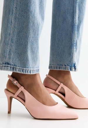 Füße tragen hellblaue Jeans und spitze rosa Slingback-Pumps mit Schnallenriemen auf weißem Boden.