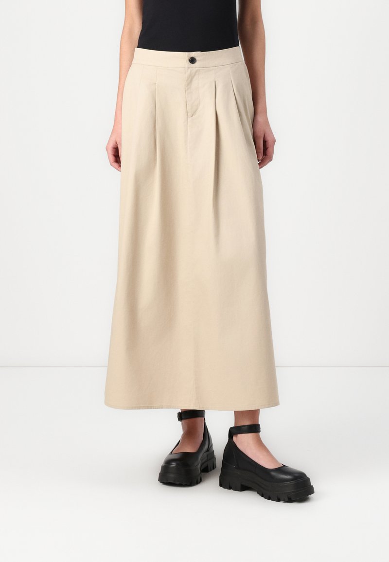 Beige midi-skjørt med folder, høy midje og frontknapper, laget av et glatt stoff. Kombinert med kraftige svarte platåsko.