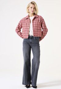 Vrouw in een rood en wit houndstoothjasje, witte top, grijze hoogwaist wijde broek en zwarte schoenen, staande tegen een witte achtergrond.