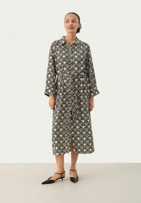 Mekko, jossa on musta ja kerma hämäräkuvio, pitkät hihat, nappisulku edessä, kaulus ja keskikokoinen helma. Yhdistetty mustiin korkokenkiin.