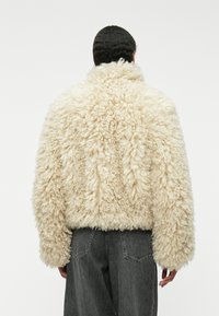 Kremowa polarowa kurtka o teksturowanej, puszystej powierzchni, w oversize'owym fasonie i miękkim kołnierzu. Noszona na ciemnoszarych spodniach.