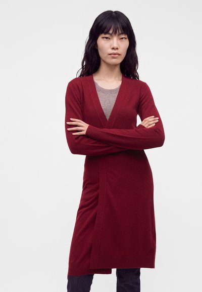 Femme aux longs cheveux foncés portant un long cardigan rouge par-dessus un haut beige, debout les bras croisés devant un fond uni.