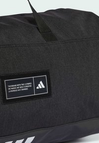 Black duffel bag with a textured fabric, featuring a rectangular label with white text and the Adidas logo on one side.