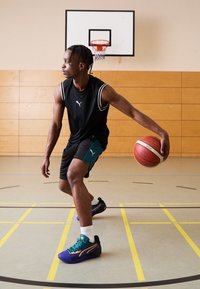 Zwarte mouwloze sportshirt, zwarte shorts met turquoise accenten, paarse en zwarte basketbalschoenen met turquoise veters, dribbelend met een basketbal.