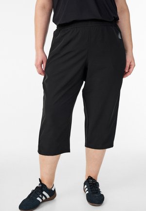 Person wearing black cropped pants, black shirt, and black sneakers with white stripes, standing on a plain white background.