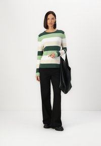 Gestreepte trui in groen, wit en beige; lange mouwen; gecombineerd met zwarte wijde broek en draagt een grote zwarte tote bag.