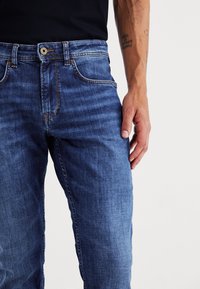 Jeans de denim en azul con un corte clásico, que presentan costuras visibles, bolsillos frontales y un cierre de botón metálico.