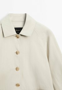 Beige knoopjack met brede kraag, vier grote knopen en zichtbare stiksels op de schouders en de kraag.