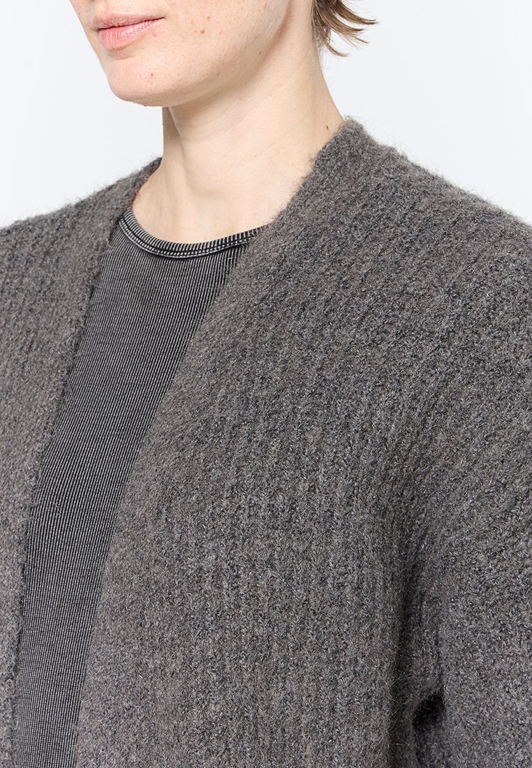 Grauer, strukturierter Cardigan mit weichem Strickfinish, offenem Vorderteil und weiten Ärmeln, kombiniert mit einem schwarz-weißen gestreiften Oberteil.