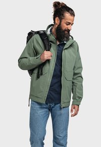 Giacca verde impermeabile con cappuccio, chiusura con zip e multiple tasche, indossata sopra una camicia blu scuro, abbinata a jeans chiari.