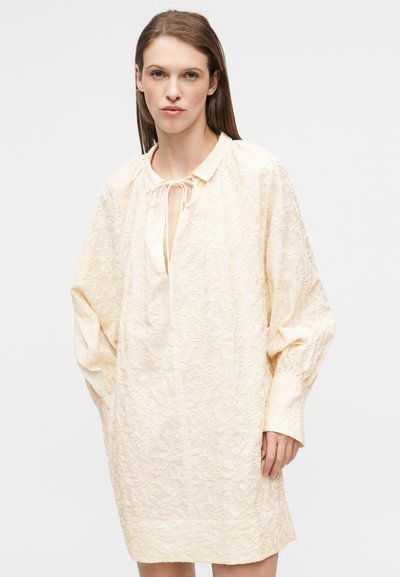 Φόρεμα με μακριά μανίκια σε ανοιχτό κρεμ ύφασμα με υφασμένο λουλουδένιο μοτίβο, με γιακά και λεπτομέρεια δεσίματος στο λαιμό.