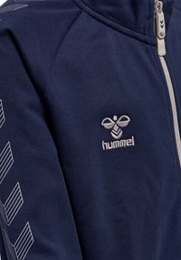 Veste zippée marine en tissu doux, ornée d'un logo Hummel blanc et d'un motif en rayures géométriques sur la manche.