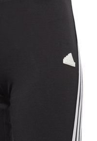 Leggings sportivi neri con una texture liscia, caratterizzati da un piccolo logo Adidas bianco vicino all'anca e tre strisce verticali bianche lungo il lato.