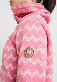 Felpa rosa con un motivo a zigzag bianco, dotata di un colletto alto, un bottone in legno e una patch rotonda in pelle sul manica sinistra.