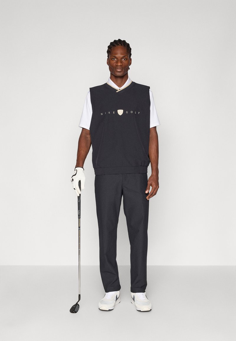 Gilet da golf senza maniche nero su una camicia bianca con colletto, pantaloni grigi, scarpe da ginnastica bianche, con un bastone da golf in mano e un guanto su una mano.