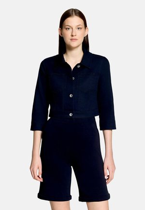 Žena nosiaca tmavomodrú krátku bundu s trojštvrťovými rukávmi a zodpovedajúce krátke nohavice po kolená, stojaca pred jednotvárnym pozadím.