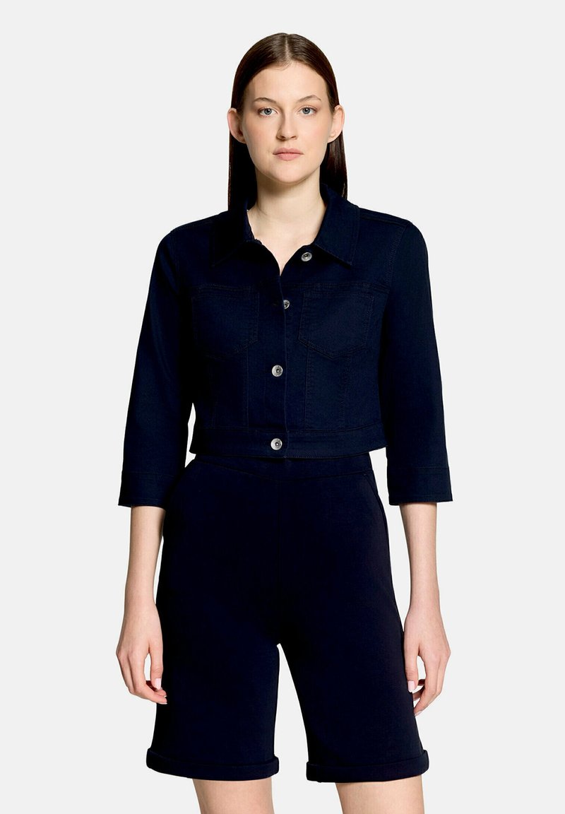 Femme portant une veste courte bleu marine foncé avec des manches trois-quarts et un short assorti longueur genou, debout devant un fond uni.