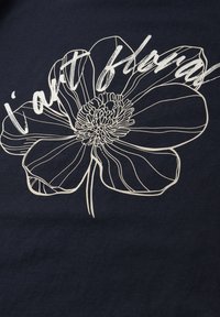 Dibujo en línea blanca de una flor grande con la frase «soy flores» escrita sobre los pétalos, sobre un fondo de tela oscura.