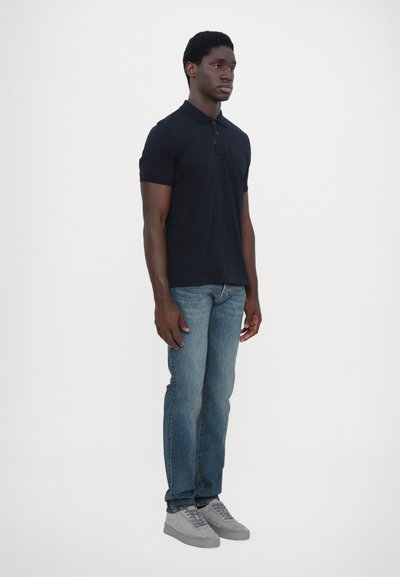 Navy polo shirt with short sleeves, paired with light blue jeans and grey sneakers. Model standing on a plain white background.