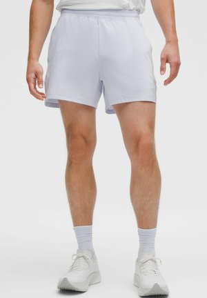 Mann trägt weiße Sportshorts, weiße Socken und weiße Laufschuhe, steht mit entspannten Händen an den Seiten.