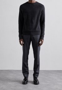 Schwarzer strukturierter Pullover mit rundem Ausschnitt, kombiniert mit schmal geschnittenen dunkelgrauen Hose und schwarzen Schnürschuhen, vor einem hellen Hintergrund.