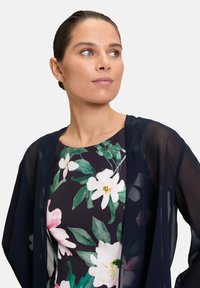 Marinefarbener transparenter Kimono über einem floral gemusterten Kleid, das weiße und rosa Blumen mit grünen Blättern vor einem dunklen Hintergrund zeigt.