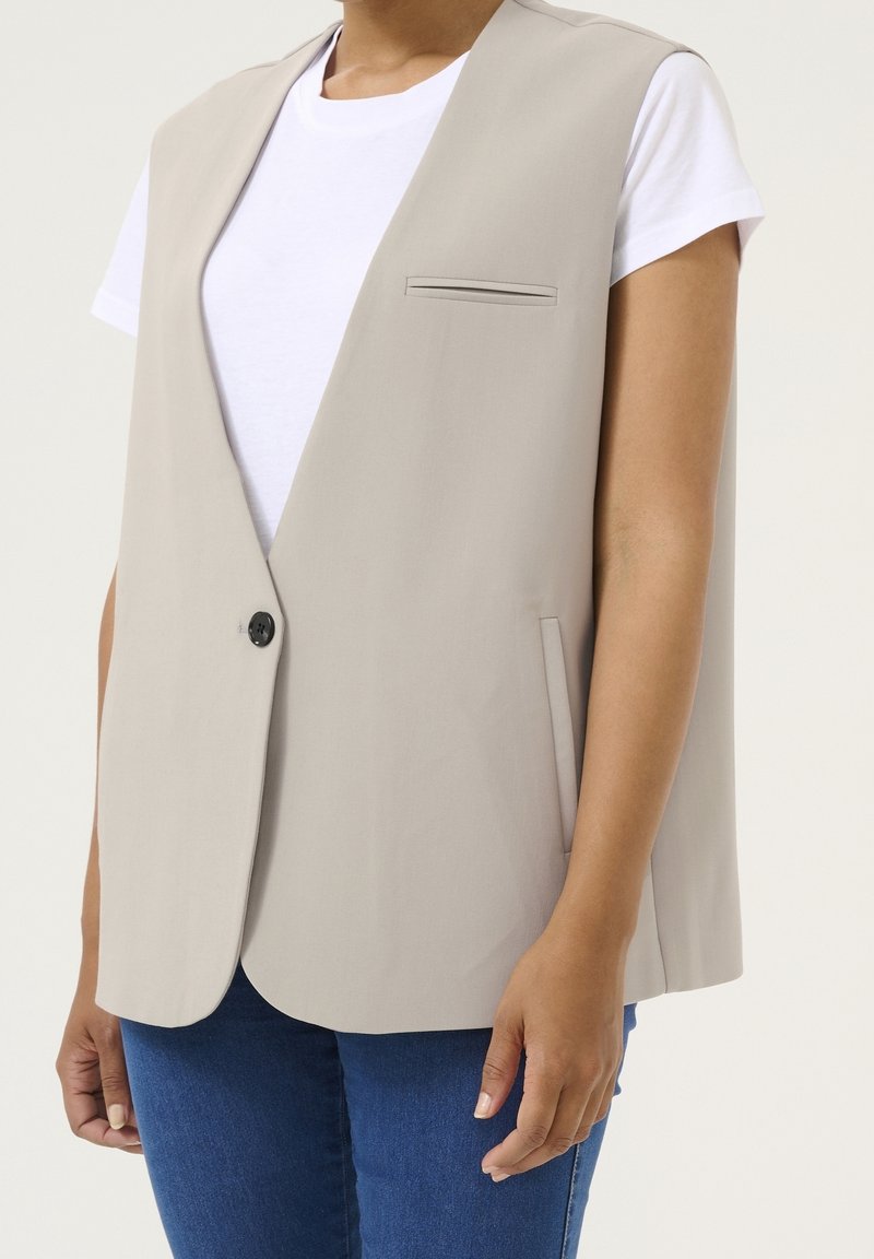 Beige ärmellose Weste mit schlanker Passform, einreihiger Knopfverschluss und einer Brusttasche. Getragen über einem weißen T-Shirt und kombiniert mit blauen Jeans.