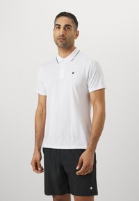 Weißes Poloshirt mit schwarzem gesticktem Logo, Kragen-Design, zwei Knopfverschluss und schwarzem Besatz. Kombiniert mit schwarzen Shorts.