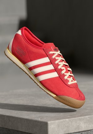Røde suede sportsko med hvide striber, snørelukning og en struktureret gummisål. Logo på siden. Klassisk retro stil.