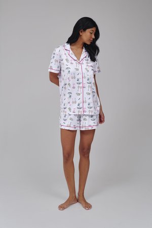 Femme debout pieds nus, portant un pyjama blanc à manches courtes avec un liseré rose et un imprimé ludique d’ustensiles et de boissons, regardant vers le bas, sur le côté.