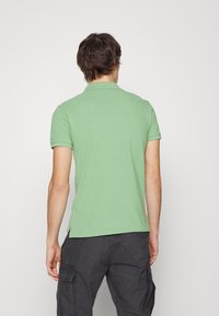 Grön bomullspolo med kort ärm, med en klassisk krage och sidoklyftor, bärs med mörka cargobyxor. Bakvy visas.