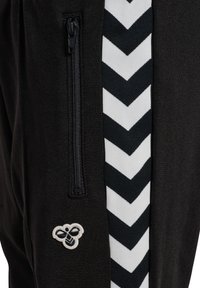 Czarna tkanina z czarną kieszenią na suwak, biało-czarny pas w jodełkę oraz małe logo pszczoły blisko dołu.