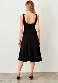 Femme aux longs cheveux blonds, debout face à un mur, portant une robe midi noire sans manches avec un dos ouvert et des sandales noires à talons.