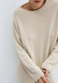 Pull côtelé beige à large encolure et manches longues, avec une coupe décontractée et une texture douce. Les mains sont placées ensemble devant.