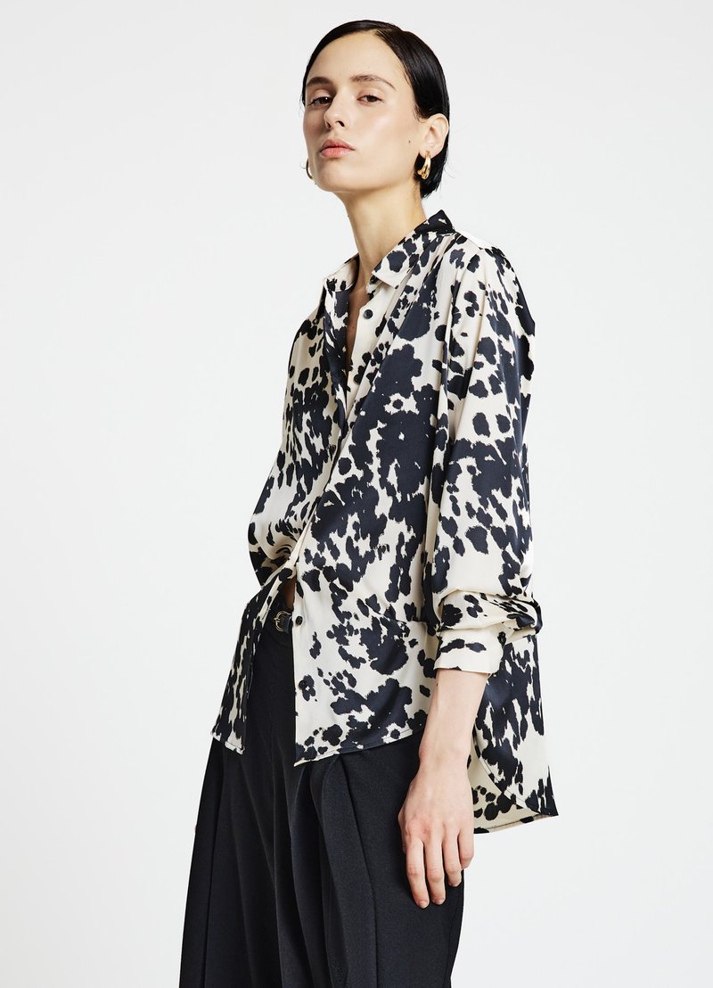 Hemdenbluse in hellem Creme mit schwarzem abstraktem Muster, mit langen Ärmeln und lockerem Schnitt, kombiniert mit hoch taillierten schwarzen Hosen.