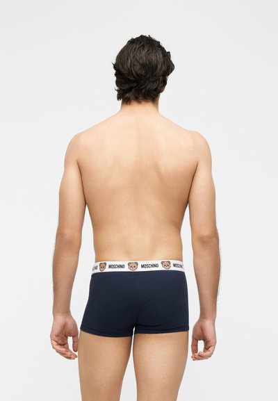 Άνδρας που φαίνεται από πίσω φορώντας σκούρα μπλε μποξεράκια με λευκή ζώνη μέσης με το λογότυπο Moschino και πρόσωπα αρκούδων teddy bear.