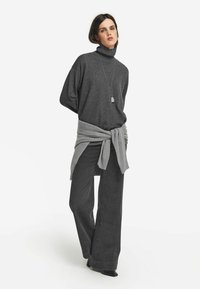 Frau in einem dunkelgrauen Rollkragenpullover, weit geschnittenen schwarzen Hosen, einem grauen Pullover um die Taille gebunden und einer langen Anhängerhalskette, die vor einem weißen Hintergrund steht.