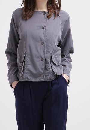 Femme portant une veste grise à boutons avec des poches à rabat et un pantalon bleu marine, les mains dans les poches, sur un fond clair uni.