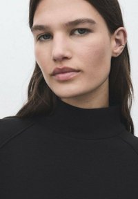 Un haut à col roulé noir avec une texture lisse, présentant des manches raglan et un col haut, porté par un mannequin ayant de longs cheveux et un arrière-plan neutre.